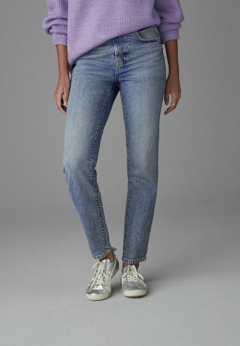Personne portant un jean en denim bleu clair, des baskets argentées et un pull lavande, debout devant un fond gris uni.