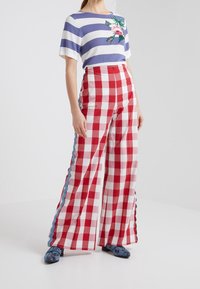 Mujer con una blusa de rayas azul y blanca con bordados de flores, pantalones de pierna ancha a cuadros rojos y blancos, y zapatos azules con un patrón.