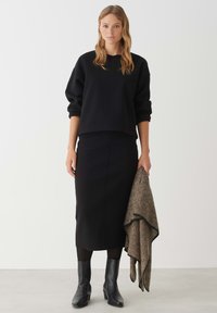 Zwarte cropped sweatshirt met verlaagde schouders, gecombineerd met een zwarte rok tot de knie. Model houdt een gestructureerde bruine sjaal vast. Zwarte enkellaarzen.