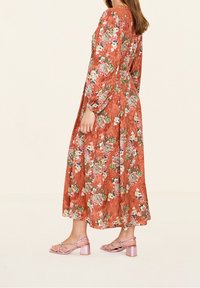 Blommig maxiklänning i rostfärg med långa ärmar, som har ett texturerat mönster och livliga blomdetaljer. Matchad med blekrosa klacksandaler.