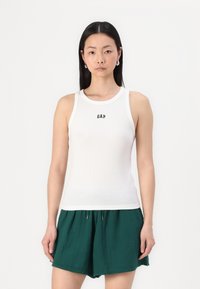Weißes, geripptes Tanktop mit rundem Ausschnitt und dem "GAP"-Logo in der Mitte, kombiniert mit grünen Shorts. Glatte Textur, figurschmeichelndes Design.