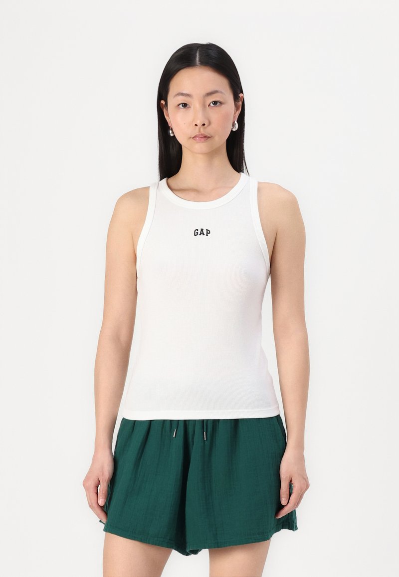 Weißes, geripptes Tanktop mit rundem Ausschnitt und dem "GAP"-Logo in der Mitte, kombiniert mit grünen Shorts. Glatte Textur, figurschmeichelndes Design.
