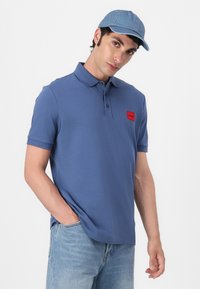 Kék póló, bordázott gallérral, rövid ujjú, piros logóval. Világoskék farmernadrággal és világoskék denim sapkával viselve.