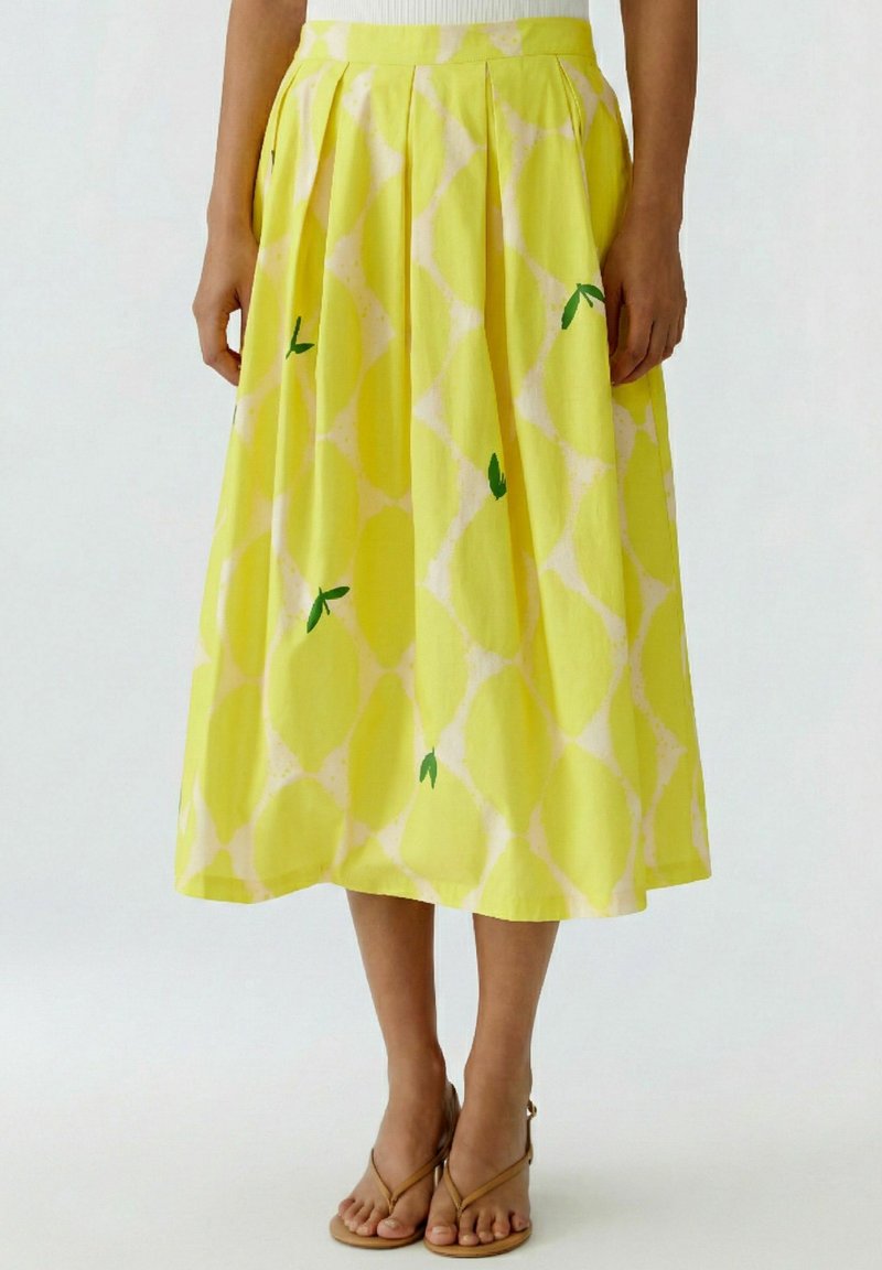 Woman wearing a pleated yellow midi skirt with a light geometric pattern and small green leaf accents, paired with beige strap sandals.