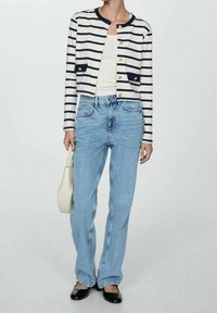 Gilet court rayé bleu marine et blanc avec des boutons dorés, porté avec un jean droit bleu clair et des ballerines noires. Tient un sac blanc.