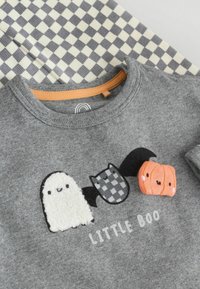 Nærbillede af gråt stof med søde patches af spøgelser, flagermus og græskar over teksten "LILLE BOO" og et grå-hvidt ternet tekstil i baggrunden.