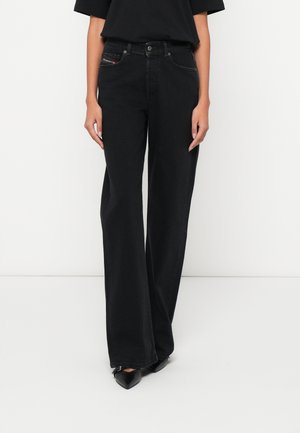 Femme portant un jean noir taille haute à jambes larges, associé à un haut noir à manches courtes et des chaussures noires à bouts pointus.