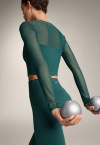 Osoba w turkusowym stroju sportowym trzymająca za plecami dwie srebrne piłki do ćwiczeń, widziana z boku i tyłu.