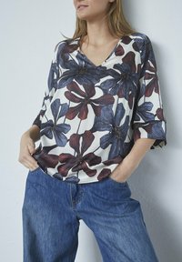 Bluse med blomsterprint, v-hals, korte ærmer, hvid baggrund, store blå og burgunder blomster, parret med blå denim jeans.