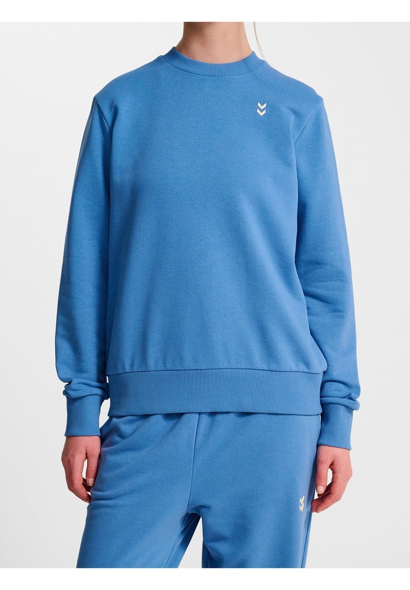 Personne portant un sweat-shirt bleu manches longues et un pantalon bleu assorti avec de petits logos blancs en forme de chevrons sur la poitrine et la cuisse.
