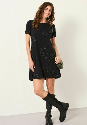 Schwarzes Kurzarmkleid mit floralem Texturdetail. A-Linie, knielang, kombiniert mit knielangen schwarzen Stiefeln und einer kleinen grünen Schultertasche.