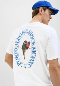 Camiseta blanca de algodón con un gráfico colorido de dos pájaros y el texto "PORTO ALEGRE SINCE MCMXC" en azul que rodea el diseño.