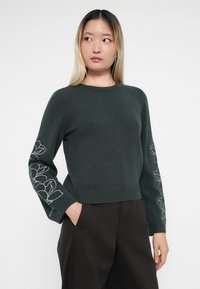 Pull vert foncé avec un col rond et ourlet côtelé. Les longues manches présentent des broderies florales blanches sur les poignets. Texture en tricot douce.