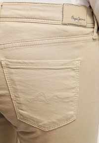 Béžové džíny s prošitými detaily na zadních kapsách a béžovou koženou nášivkou loga Pepe Jeans London na pase.