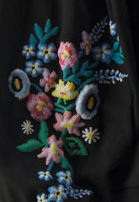 Svart stoff med fargerik blomsterbroderi i rosa, blått, gult og grønt. Detaljert søm fremhever ulike blomsterformer.