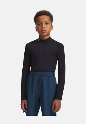 Junge steht mit Blick nach vorne, trägt ein eng anliegendes schwarzes Langarmshirt und lockere blaue Shorts vor einem einfarbigen weißen Hintergrund.