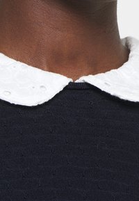 Marinblått textilier med en vit broderad krage som har en scalloped kant. Närbild visar kragen mot jämn hud.