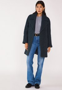 Cappotto in misto lana verde scuro con ampi rever, abbinato a un maglione grigio chiaro e jeans a zampa d'elefante blu. Scarpe nere con finitura lucida.