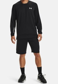 Felpa sportiva nera con collo a giro, maniche lunghe e logo Under Armour. Abbinata a pantaloni corti neri e scarpe da corsa.