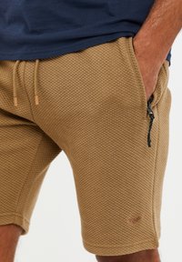 Homme portant un short marron texturé avec une taille à cordon de serrage et une poche zippée latérale, la main reposant dans la poche, associé à une chemise marine.