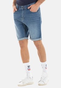 Shorts en denim à lavage clair, avec des revers, un design à cinq poches et une fermeture par bouton à l'avant. Portés avec des chaussettes blanches et des baskets.
