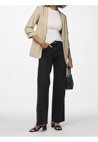 Femme portant un blazer beige, un haut blanc, un jean noir à jambes larges, des sandales à talons noires, tenant un sac à main noir, une main dans la poche.