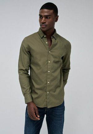 Camisa de botones verde oliva con un patrón texturizado, de mangas largas y un corte relajado, combinada con jeans azul oscuro.