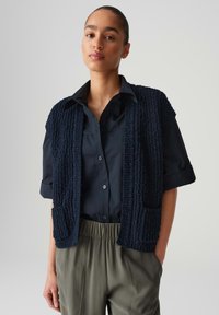 Gilet blu navy a trama con apertura frontale, dotato di due tasche a toppa, indossato sopra una maglietta scura. Modello in pantaloni larghi grigio chiaro.