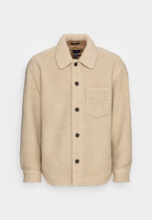 Beige sherpa fleecejakke med sorte knapper, en brystlomme på venstre side og en afslappet krave, vist på en hvid baggrund.
