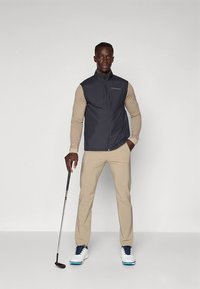 Tamsiai pilka užtraukiama liemenė dėvima ant smėlio spalvos ilgomis rankovėmis marškinio, derinami smėlio spalvos kelnės ir balti golfo batai. Laiko golfo lazdą. Paprastas dizainas.