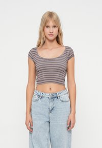 Ung kvinne med langt blondt hår har på seg en stripete crop top og lyseblå høyt liv jeans foran en ensfarget hvit bakgrunn.