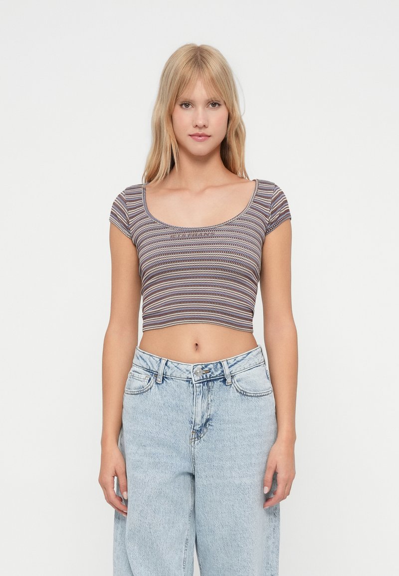 Ung kvinne med langt blondt hår har på seg en stripete crop top og lyseblå høyt liv jeans foran en ensfarget hvit bakgrunn.