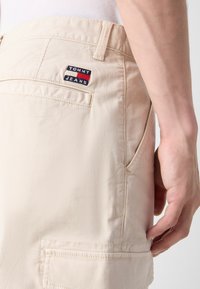 Bermudas beige de corte cargo fabricadas en tejido ligero, con un bolsillo trasero que muestra un parche con el logo "TOMMY JEANS" en azul marino, rojo y blanco.