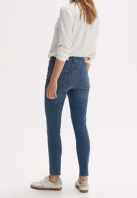 Úzké džíny v tmavě modrém denimu s vysokým pasem, vybavené zadními kapsami a našitou cedulkou na pásku, doplněné bílými teniskami.