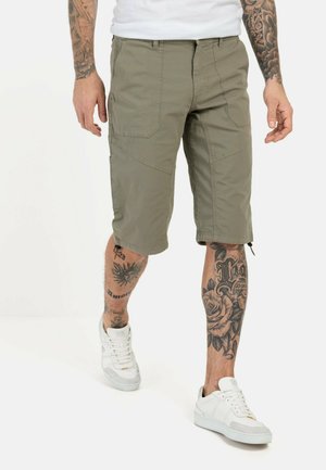 Khaki cargo shorts med flere lommer, lige snit og justerbare manchetter. Bæres med hvide sneakers og viser tatuerede ben.