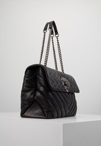 Černá prošívaná kožená kabelka s klopou, řetízkovými popruhy a ozdobným kovovým emblémem na přední straně. Hladký povrch a pevný tvar.