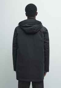 Czarna parka z kapturem o gładkim, matowym wykończeniu. Charakteryzuje się wydłużonym krojem, bocznymi kieszeniami oraz regulowanym kapturem, zaprojektowana z myślą o ochronie przed zimnem.