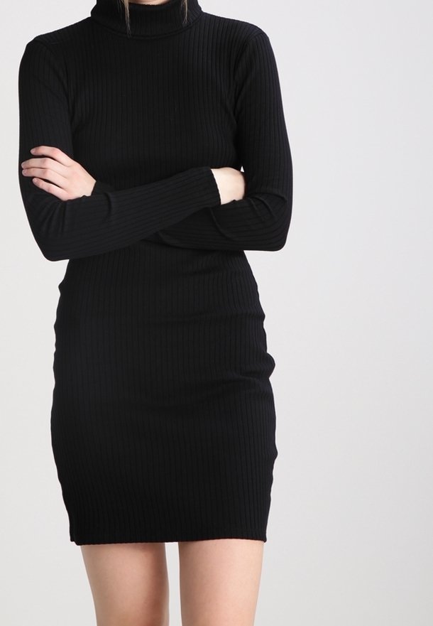 Femme portant une robe noire côtelée ajustée à manches longues et col roulé, bras croisés devant un fond clair uni.