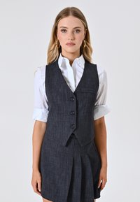 Gilet in lino blu scuro con cinque bottoni, indossato sopra una camicia bianca. Presenta una piccola tasca e un fondo svasato, adatto per contesti formali.