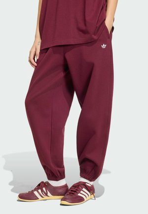 Person iført bourgogne Adidas joggers og matchende sneakers med hvide striber, stående med hænderne i lommerne mod en ensfarvet baggrund.