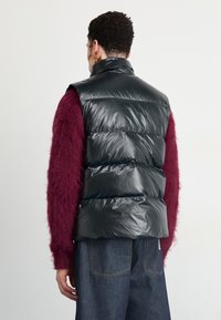 Man i en glänsande svart pufferväst över en bordeauxfärgad fluffig tröja och mörkblå denimjeans, vänd bort från kameran mot en enkel vit bakgrund.