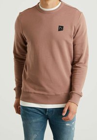 Sudadera de cuello redondo de manga larga en color rosa desvaído, hecha de tela texturizada, con un pequeño parche de logo negro en el pecho y puños de canalé.