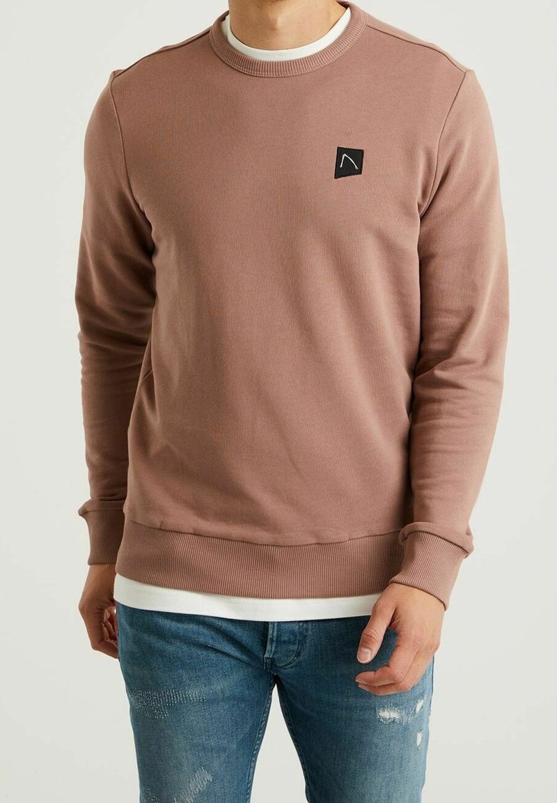 Sudadera de cuello redondo de manga larga en color rosa desvaído, hecha de tela texturizada, con un pequeño parche de logo negro en el pecho y puños de canalé.