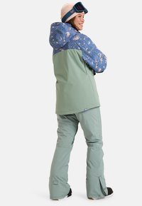 Green and blue snowboard jacket with floral print, paired with matching green pants, worn by a person with a pink hat and goggles.