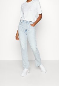 Světle modré džíny s vysokým pasem a rovnými nohavicemi, nošené s bílým tričkem s drobným grafickým designem a bílými teniskami.