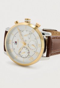 Ανδρικό ρολόι χειρός με χρυσή στεφάνη, λευκό καντράν με ρωμαϊκούς αριθμούς, τρεις υποδείκτες για τη μέρα, την ημερομηνία και 24 ώρες, καθώς και καφέ δερμάτινο λουράκι.