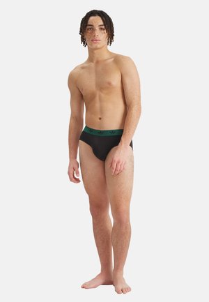 Nuori mies, jolla on pisamainen iho ja rastat, pukeutuneena mustiin alushousuihin, joissa vihreä vyötärönauha, seisoo paljain jaloin valkoista seinää vasten.