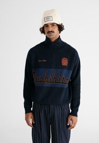 Granatowy sweter z wysokim kołnierzem, zamek z przodu oraz tekstem "Prohibite" w kontrastujących złotych i niebieskich kolorach. Noszony z pasiastymi spodniami i kremową dzianinową czapką.