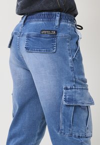 Világoskék farmer cargo nadrág, rugalmas derékkal, oldalsó zsebekkel és hátsó foltlogóval. Texturált, laza szabású dizájn.