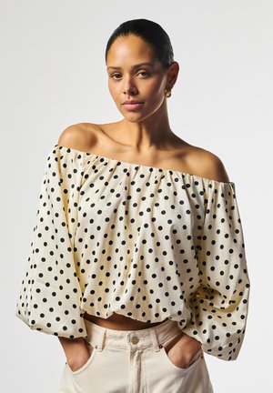 Vrouw met achterovergekamd haar, gekleed in een witte off-shoulder blouse met zwarte polkadots en lichtbeige broek, handen in de zakken.